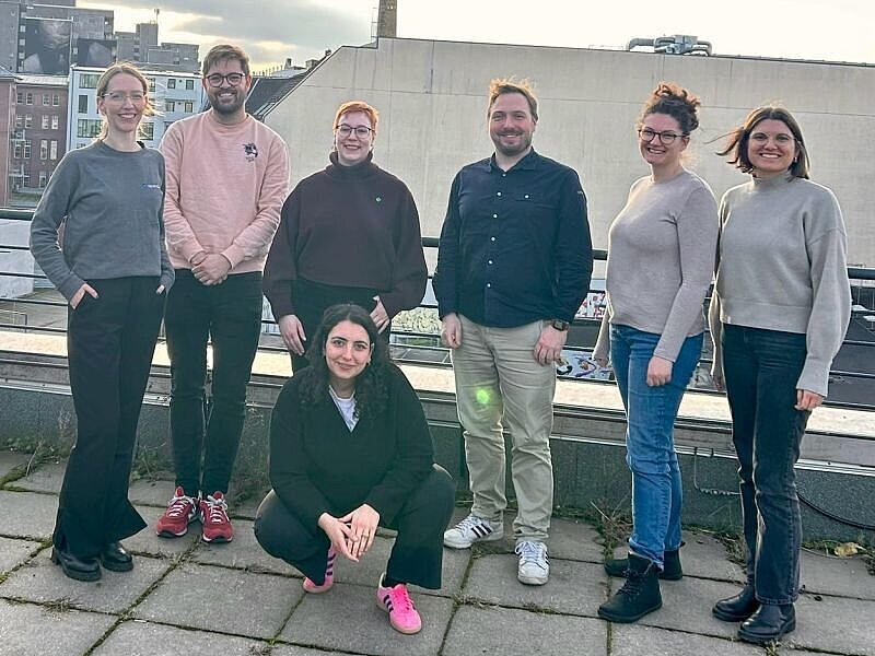 Sieben Personen bei Sonnenschein auf einer Dachterasse, im Hintergrund Gebäude.